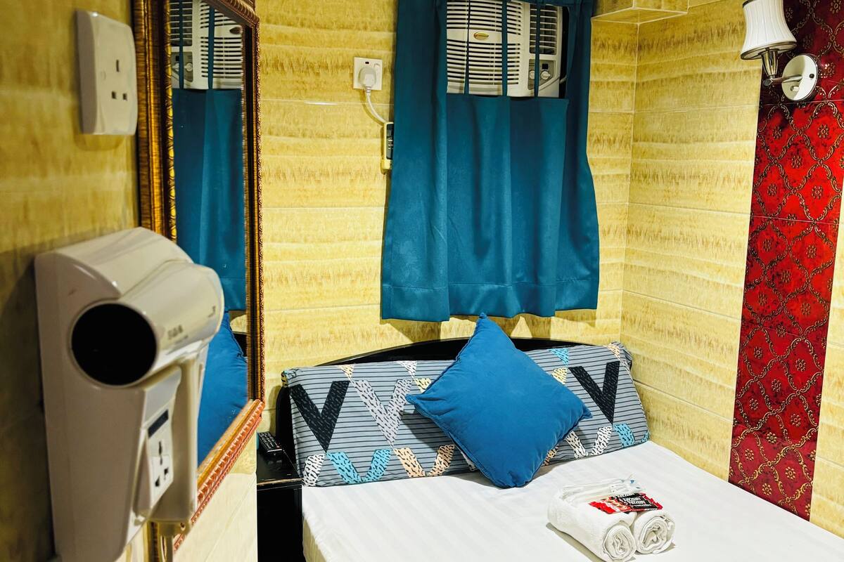 A hotel room with a bed, a hairdryer, a window with curtains, a wall-mounted lamp, and a patterned wall.