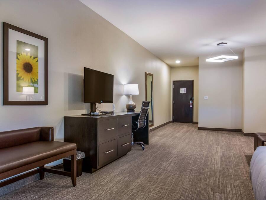 Une chambre d’hôtel avec un lit, un bureau avec un ordinateur, une chaise, une lampe, un téléviseur, un tableau encadré et une porte.
