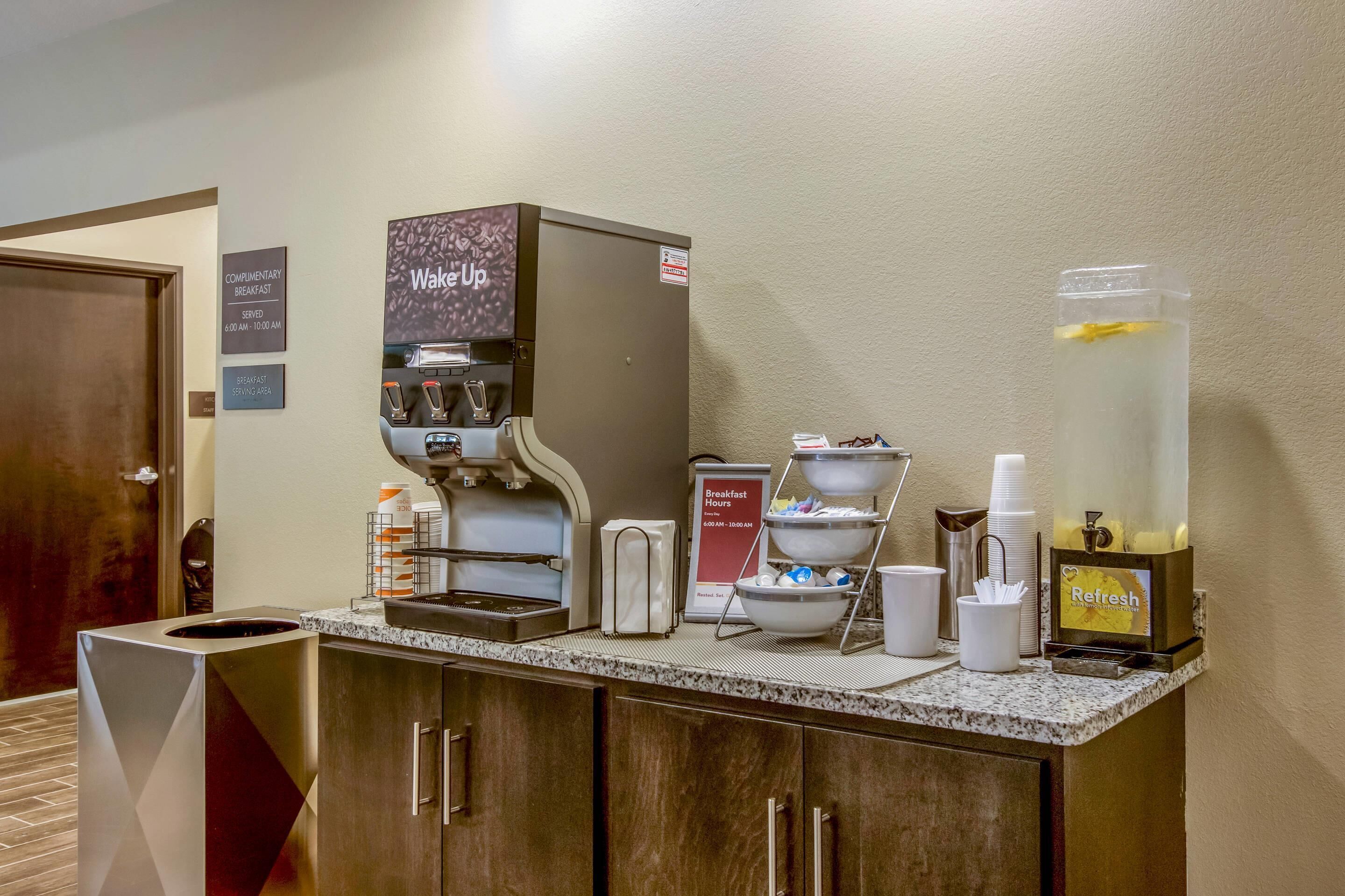 A breakfast area with a coffee machine, a water dispenser, and a sign indicating complimentary breakfast hours.