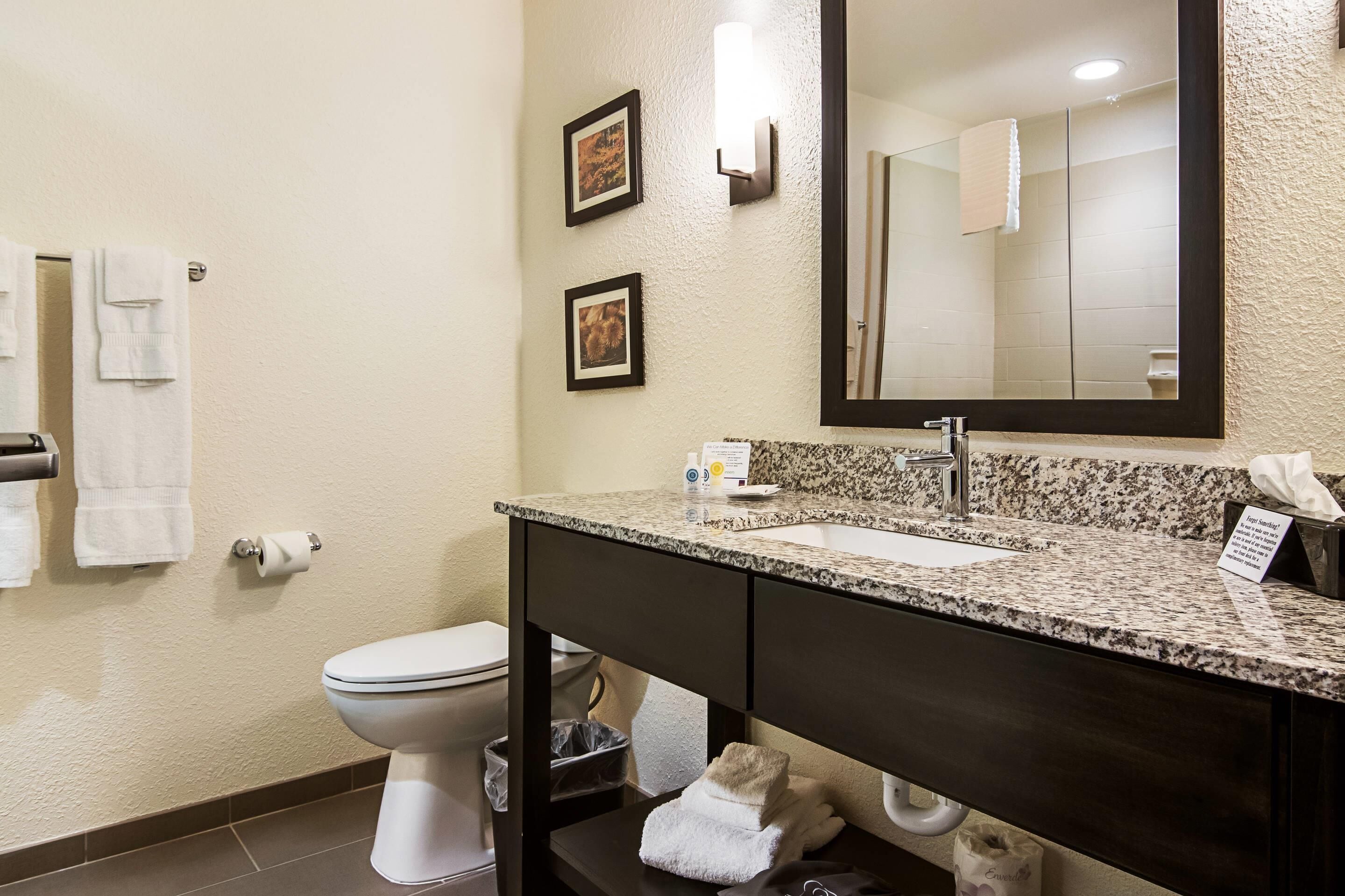A bathroom with a toilet, sink, and mirror. There are towels on the wall and a trash can.