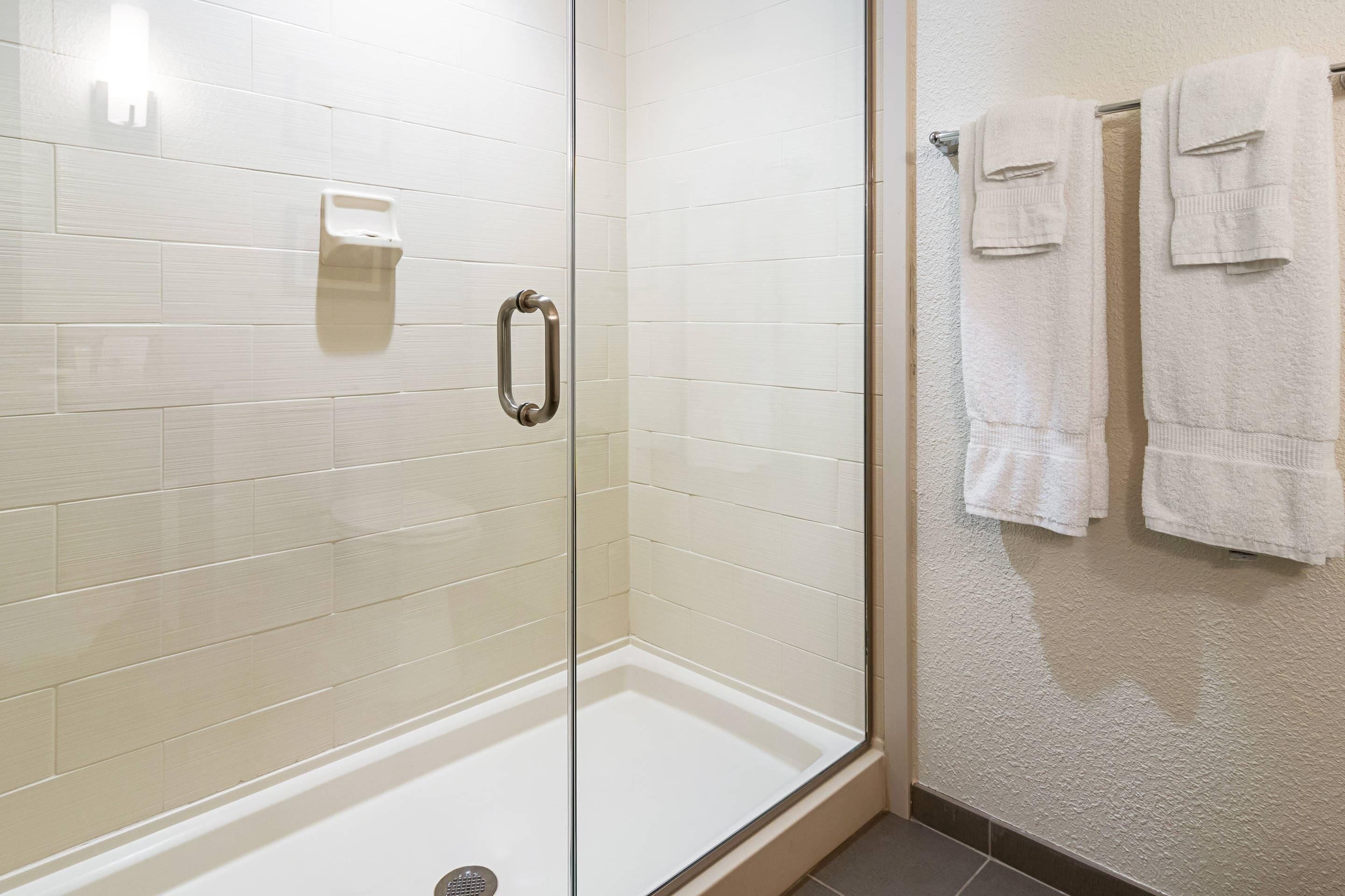 A glass-enclosed shower with a white tiled wall, a handrail, and a towel rack with two towels.