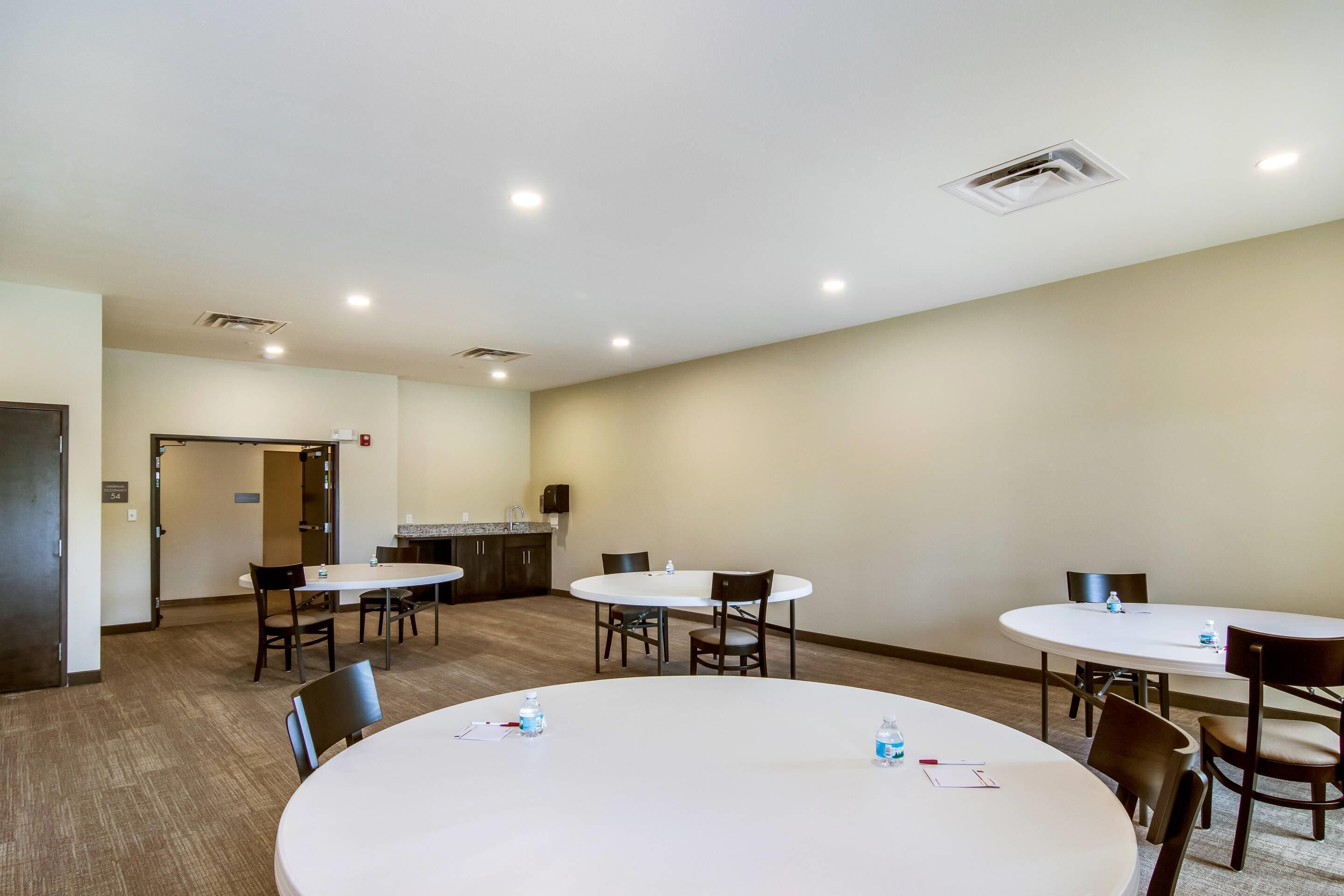 A conference room with four round tables, each with chairs, and a kitchenette area in the background.
