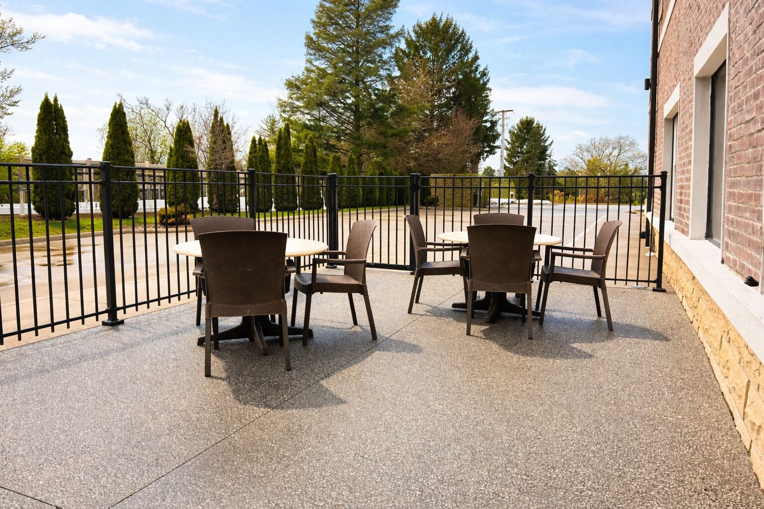 An outdoor patio with four wicker chairs and a table, surrounded by a black metal fence, overlooking a parking area and trees.