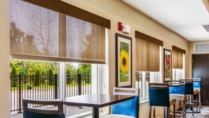 Habitación de hotel con balcón, un ventanal grande con vistas a la vegetación, un cuadro de girasol enmarco y una zona de bar con taburetes y una mesa.