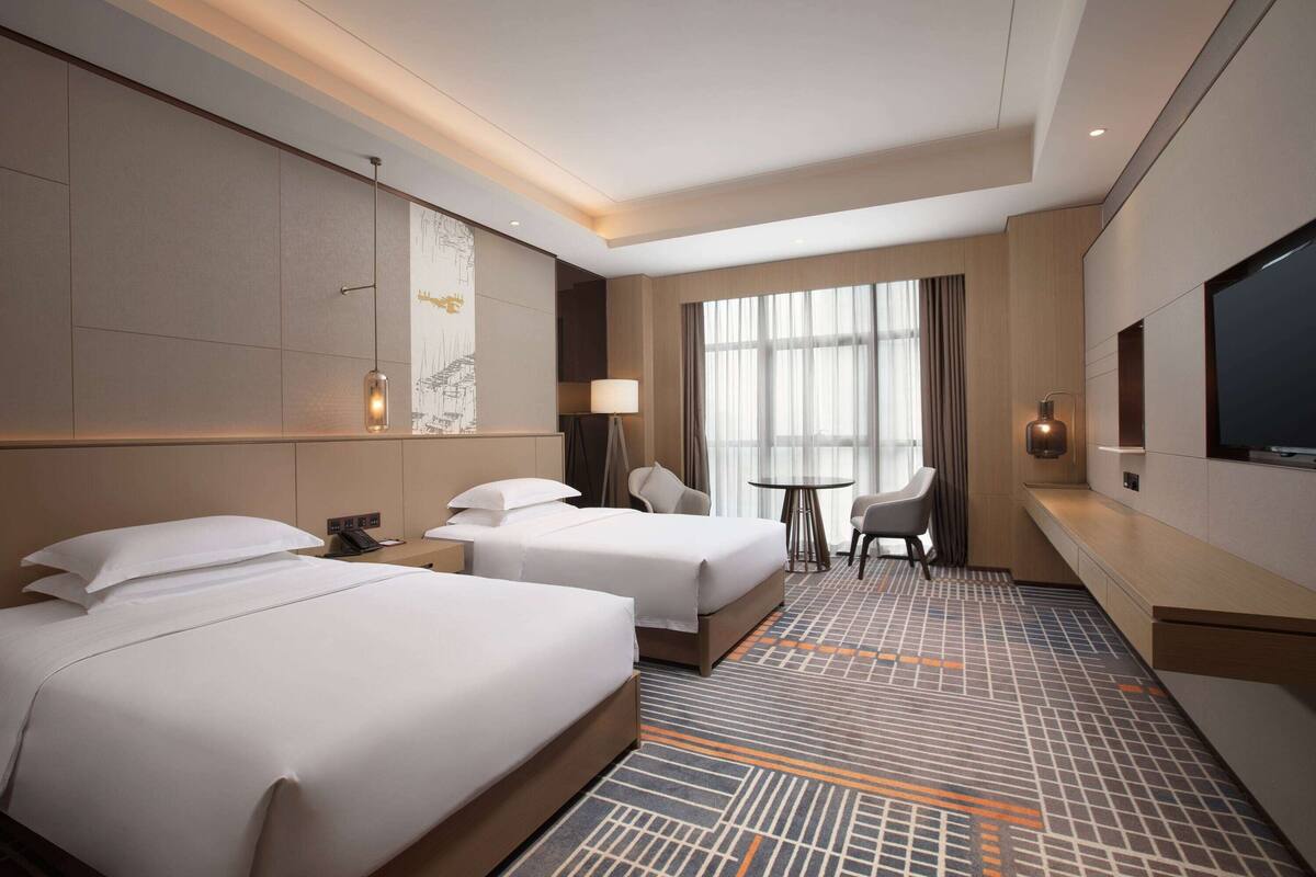 Een hotelkamer met twee bedden, een bureau, een stoel, een televisie en een groot raam met gordijnen.