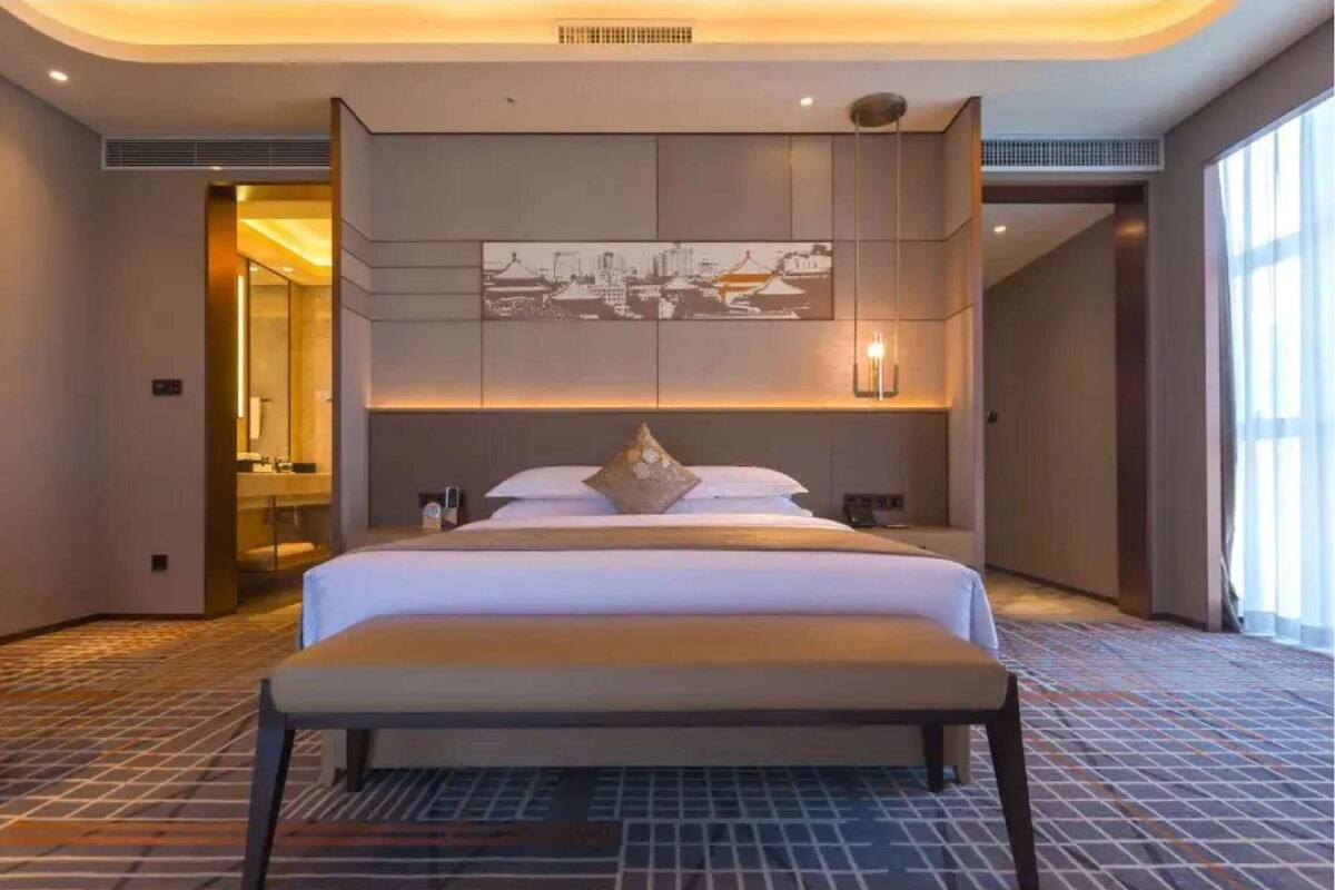 Een moderne hotelkamer met een groot bed, een bankje, een stadslandschap op de muur en een groot raam voor natuurlijk licht.