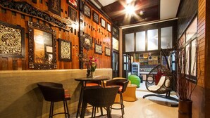 Uma área de bar com cadeiras de vime, uma pequena mesa redonda com um vaso de flores e uma parede decorativa com obras de arte emolduradas.