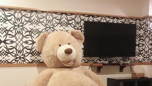 A large teddy bear sitting on a cushioned bench in a room with patterned wallpaper and a flat-screen TV mounted on the wall. - Star B&B (Tainan)