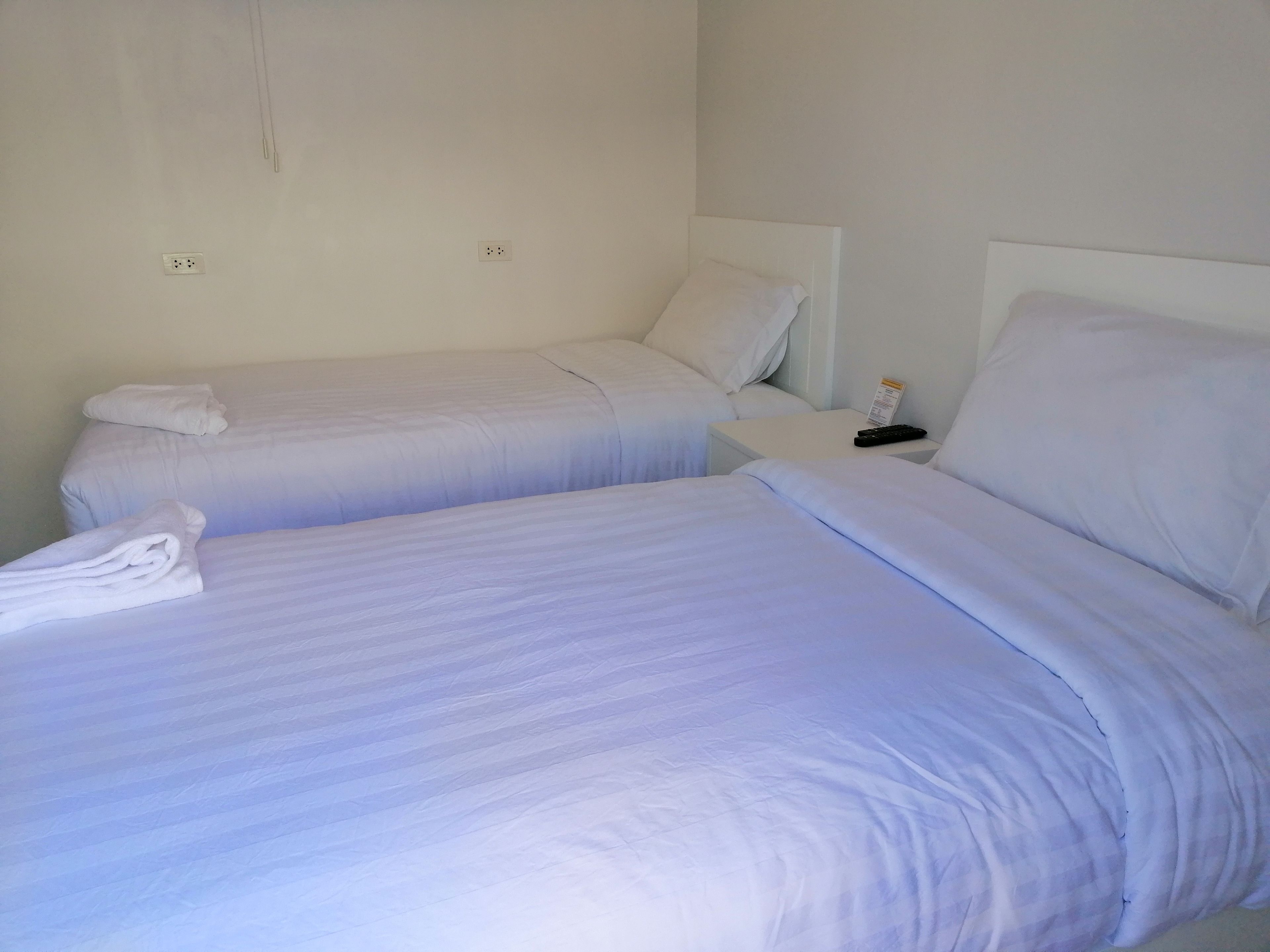 Deux lits jumeaux avec du linge de lit blanc, une table de chevet avec une télécommande et une prise électrique au mur.