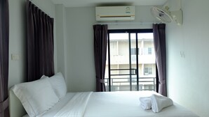 Habitación de hotel con cama, ropa de cama blanca, aire acondicionado, ventilador de techo y vistas a un edificio a través de una ventana con cortinas.