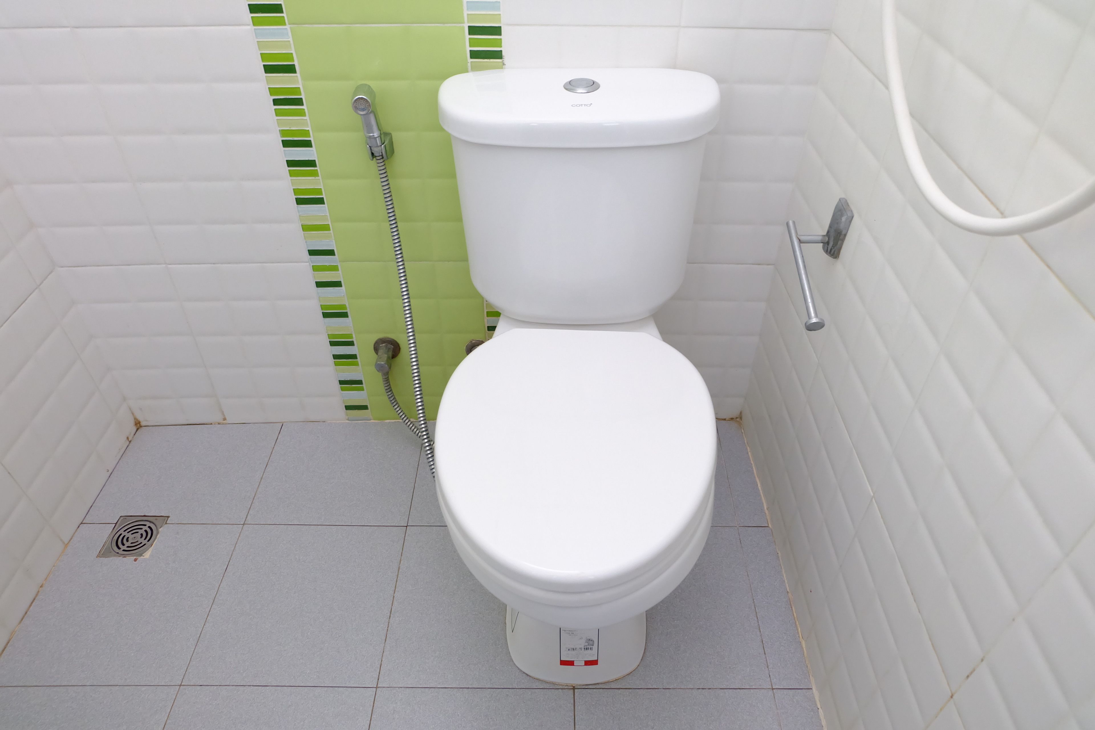 A white toilet with a green and white tiled wall, a metal handrail, and a visible drain.