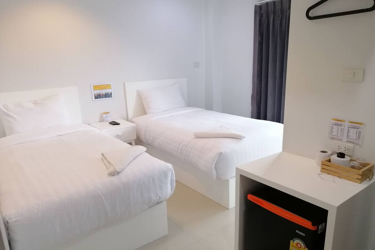 Habitación de hotel con dos camas, una mesita de noche pequeña, una mesita auxiliar de madera, un cubo de basura y un tablón informativo en la pared.