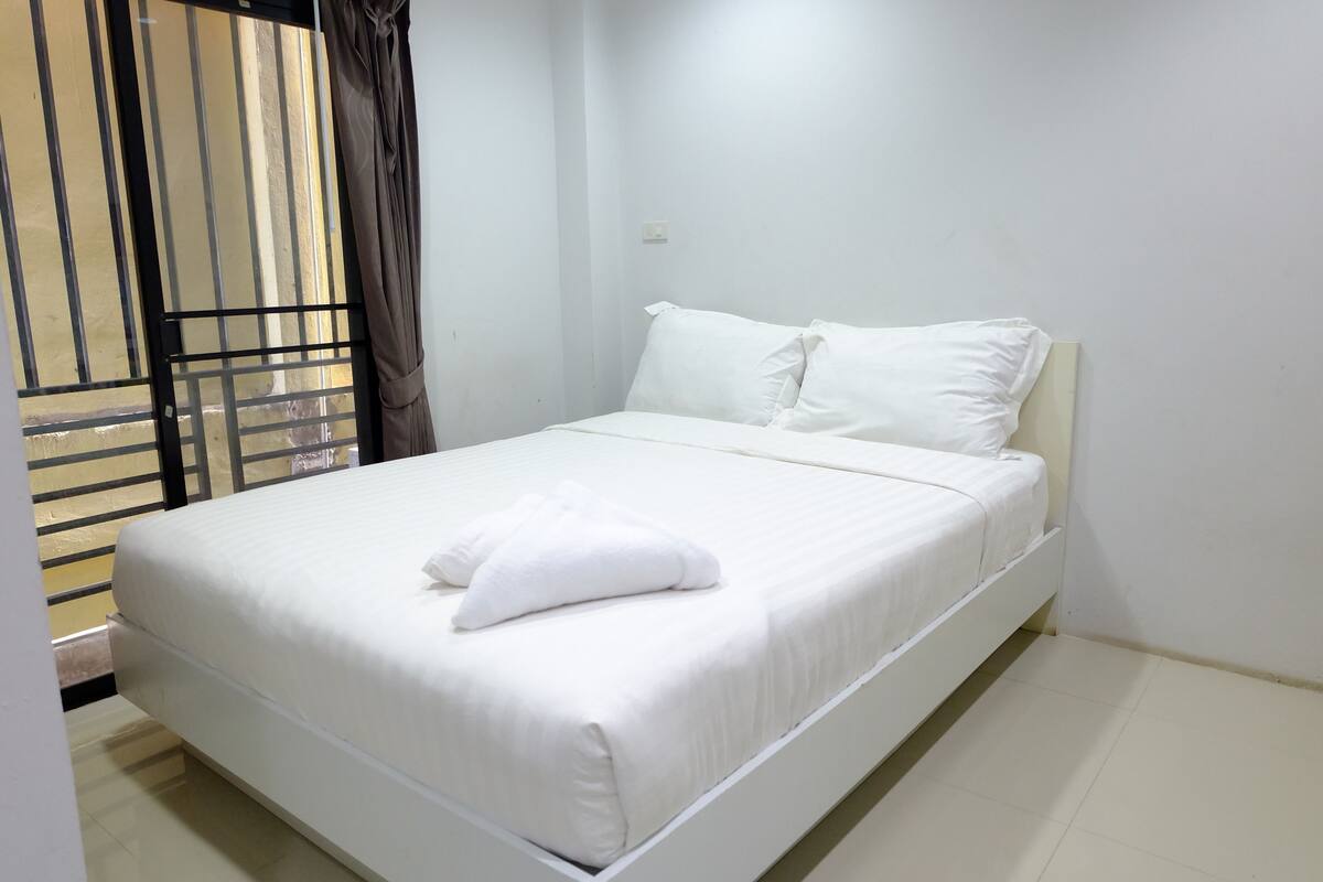 Una cama bien hecha con ropa de cama y almohadas blancas, una ventana con persianas y una pared blanca lisa.