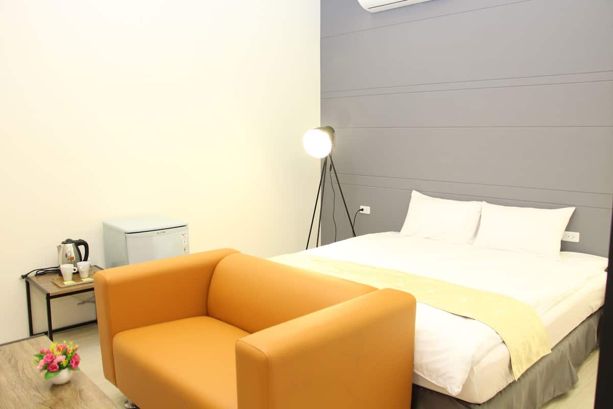A hotel room with a bed, a small table with a coffee maker, a chair, and a bedside lamp.