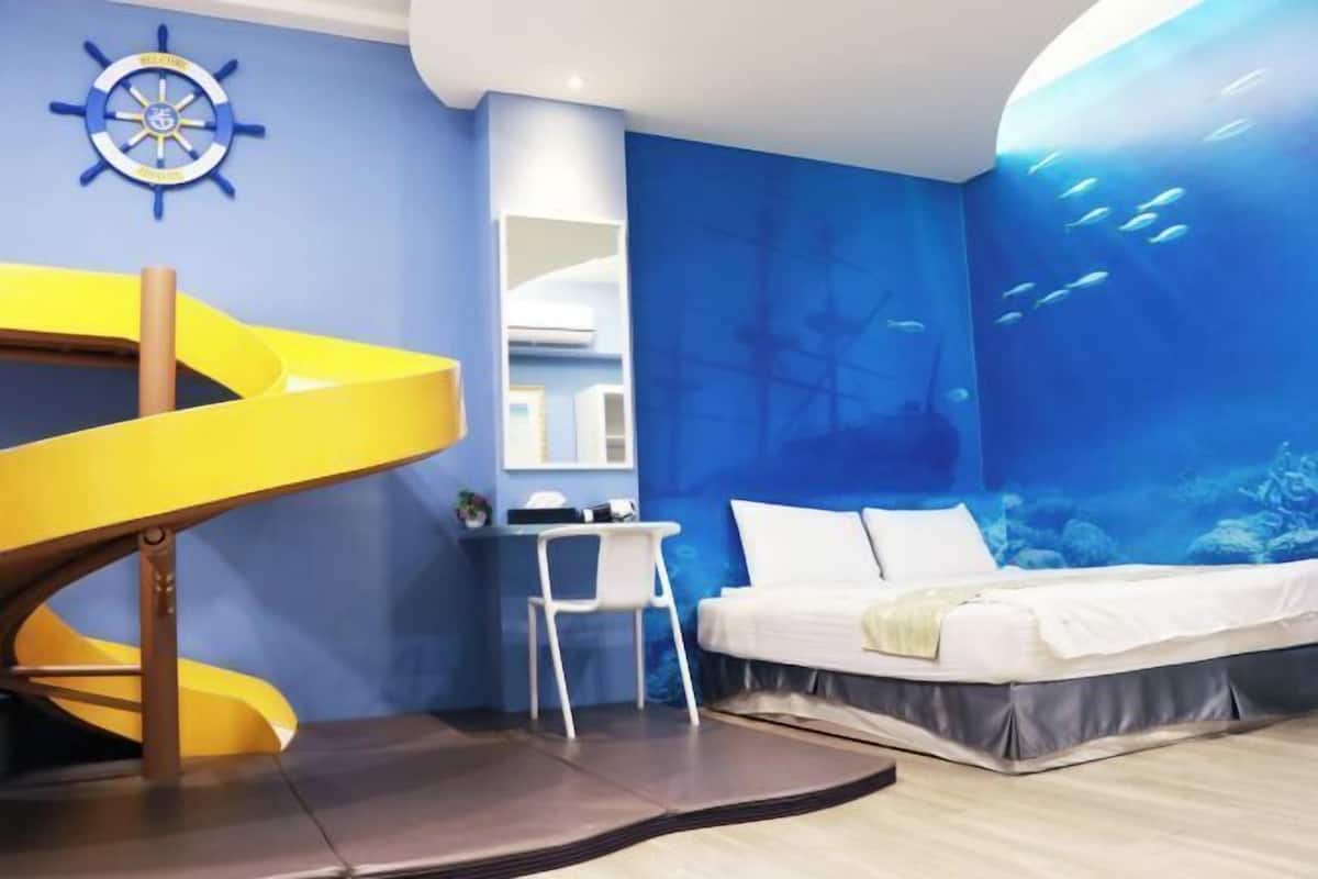 A room with a bed, a desk, a chair, a yellow spiral slide, and a wall mural of an underwater scene.