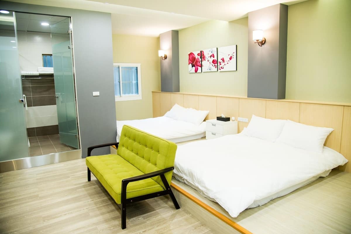 A hotel room with two beds, a green chair, a nightstand, and a painting on the wall.