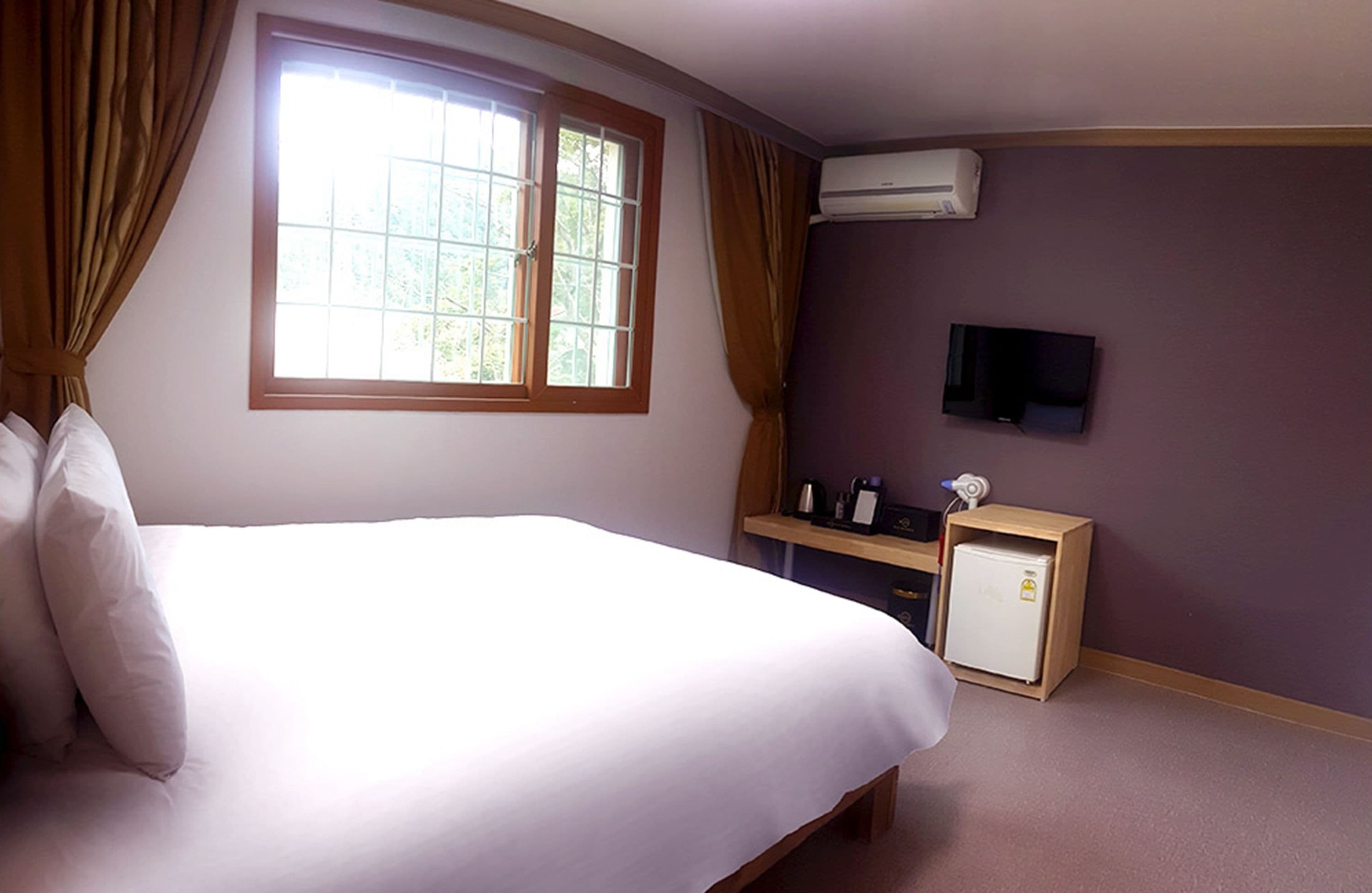 A bedroom with a bed, a window with curtains, a television, and a small refrigerator.