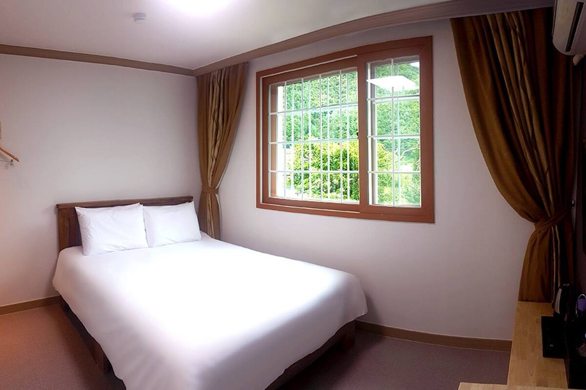 A hotel room with a bed, a window with curtains, a wall-mounted air conditioner, and a wooden floor.