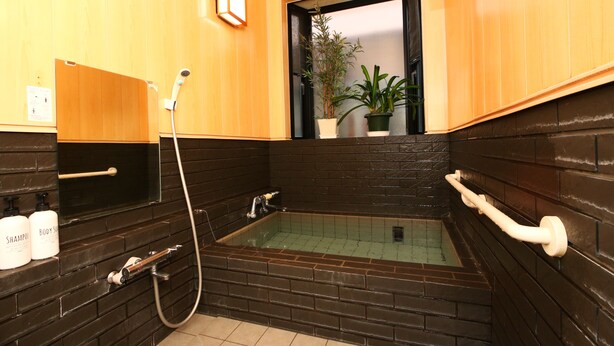 Ein Badezimmer mit einer schwarzen Eckbadewanne, einem Holzkosmetikschrank mit zwei Shampoo-Flaschen und einem Topf mit Pflanze auf der Fensterbank.