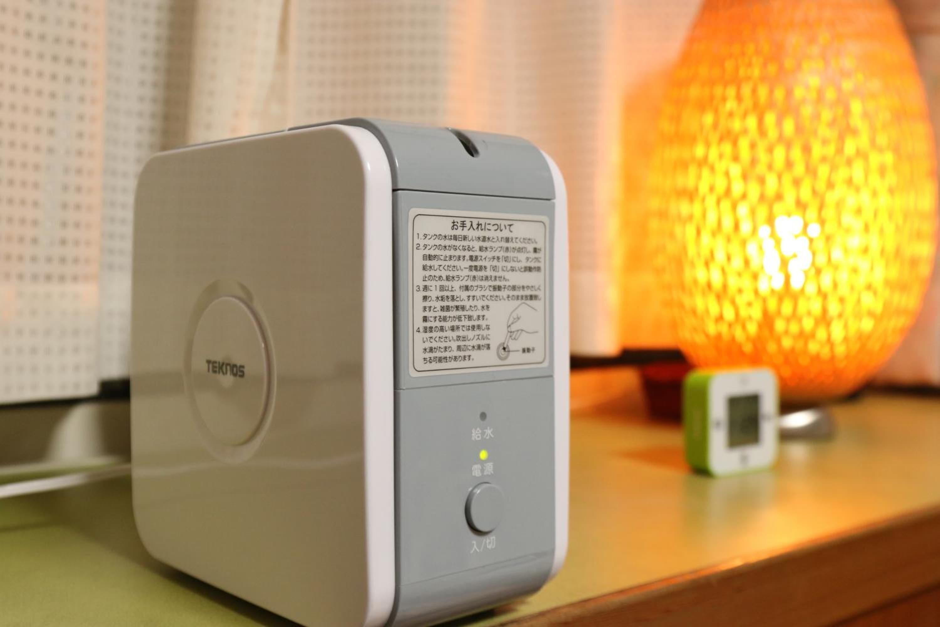 A white TEKNO5 device with Japanese text on its side, placed on a surface with a small green device and a glowing orange spherical object in the background.
