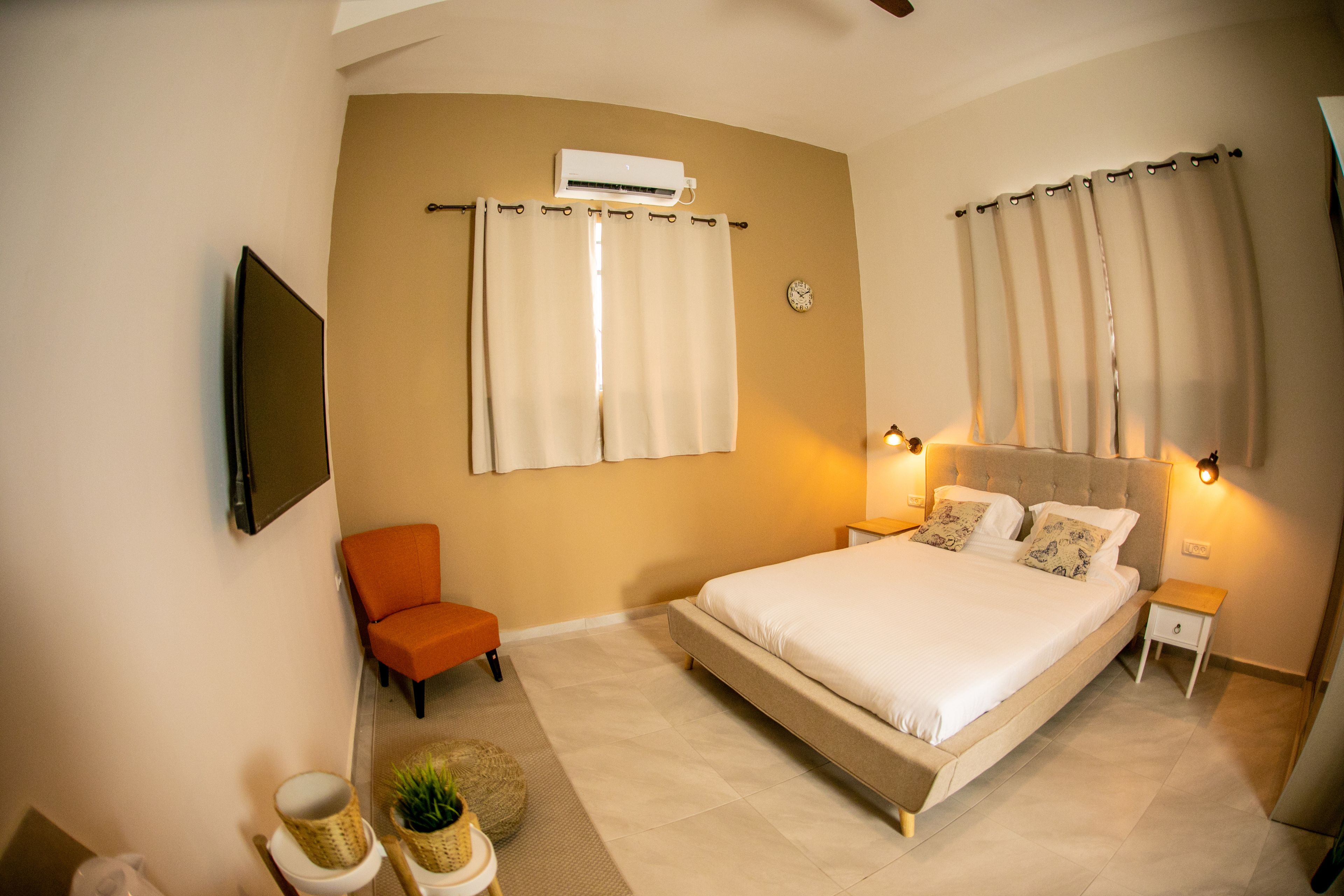 Un dormitorio con una cama grande, una silla naranja, una mesita con una planta y un equipo de aire acondicionado en la pared.