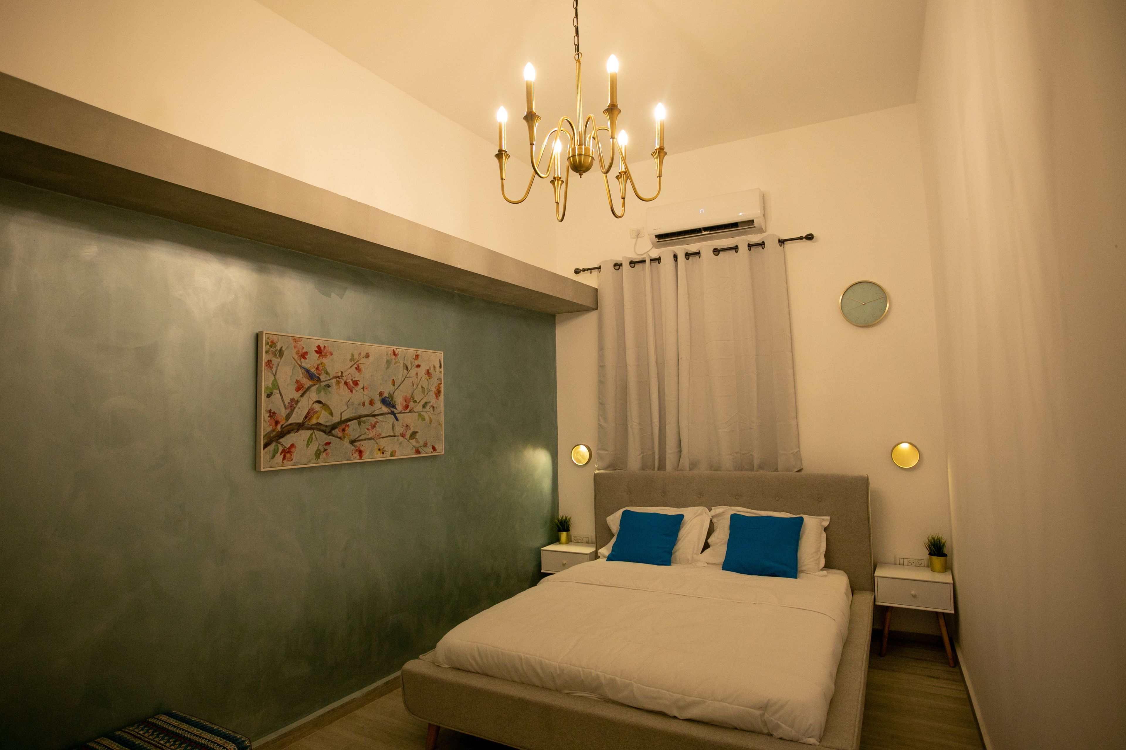 A bedroom with a bed, a nightstand, a wall-mounted clock, a chandelier, and a painting.