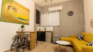 A compact living space with a kitchenette, a yellow sofa, a dining area with stools, and a large painting on the wall.