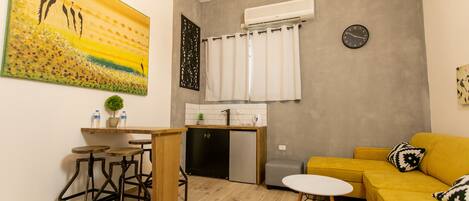 A compact living space with a kitchenette, a yellow sofa, a dining area with stools, and a large painting on the wall.
