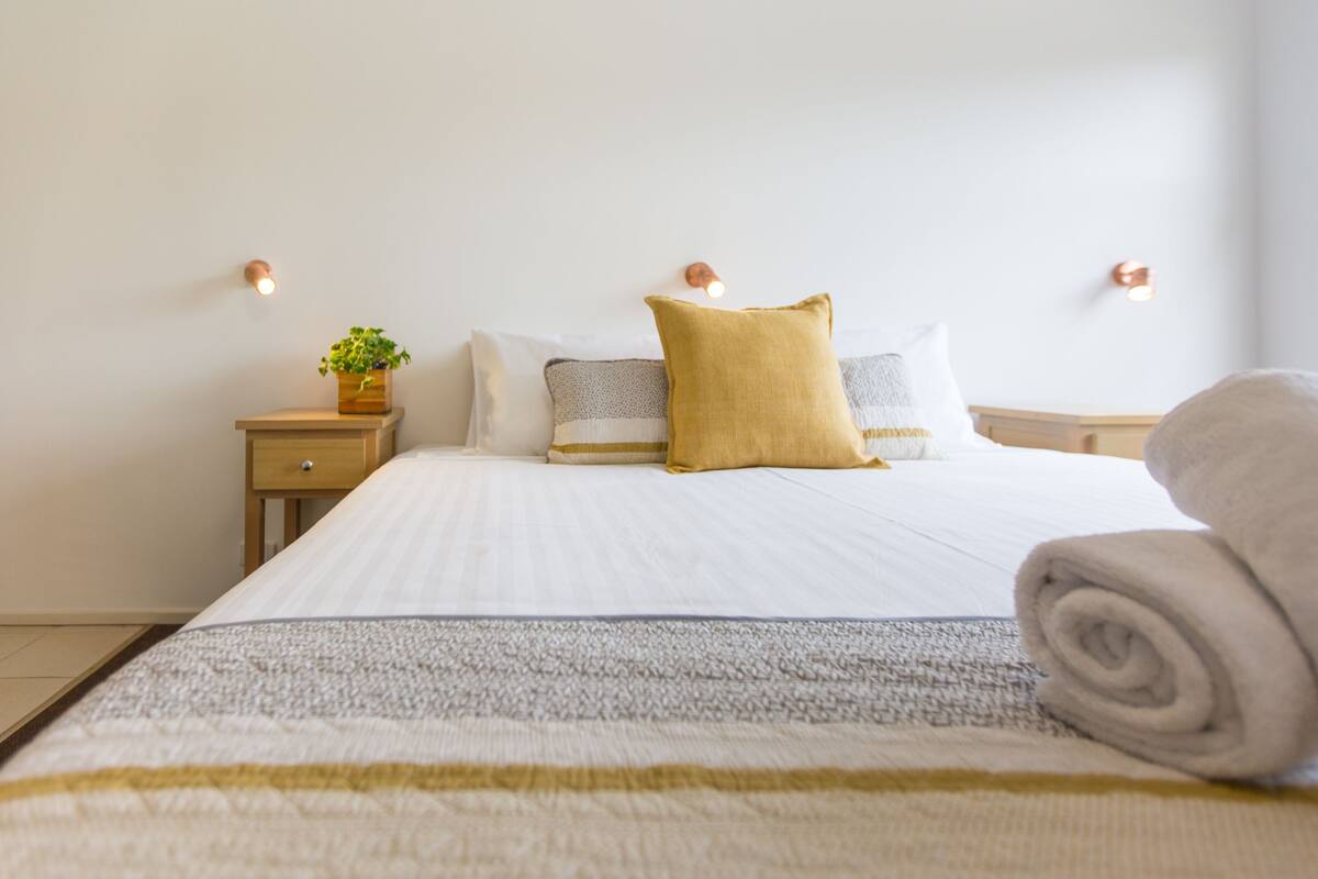 A neatly made bed with a rolled towel, a bedside plant, and a small wooden nightstand.