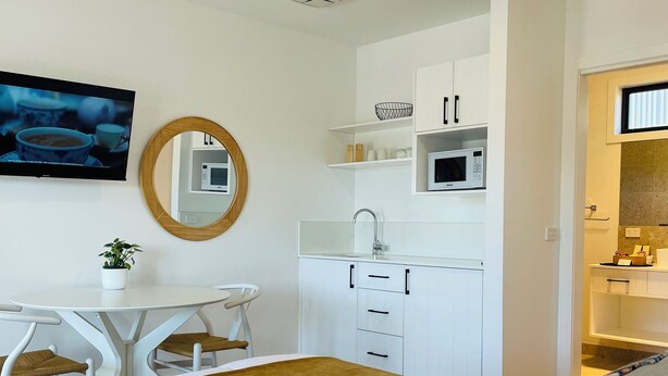 Een moderne slaapkamer met een bed, bureau, stoel, plafondventilator, televisie en een keukenhoek met kasten en een magnetron.