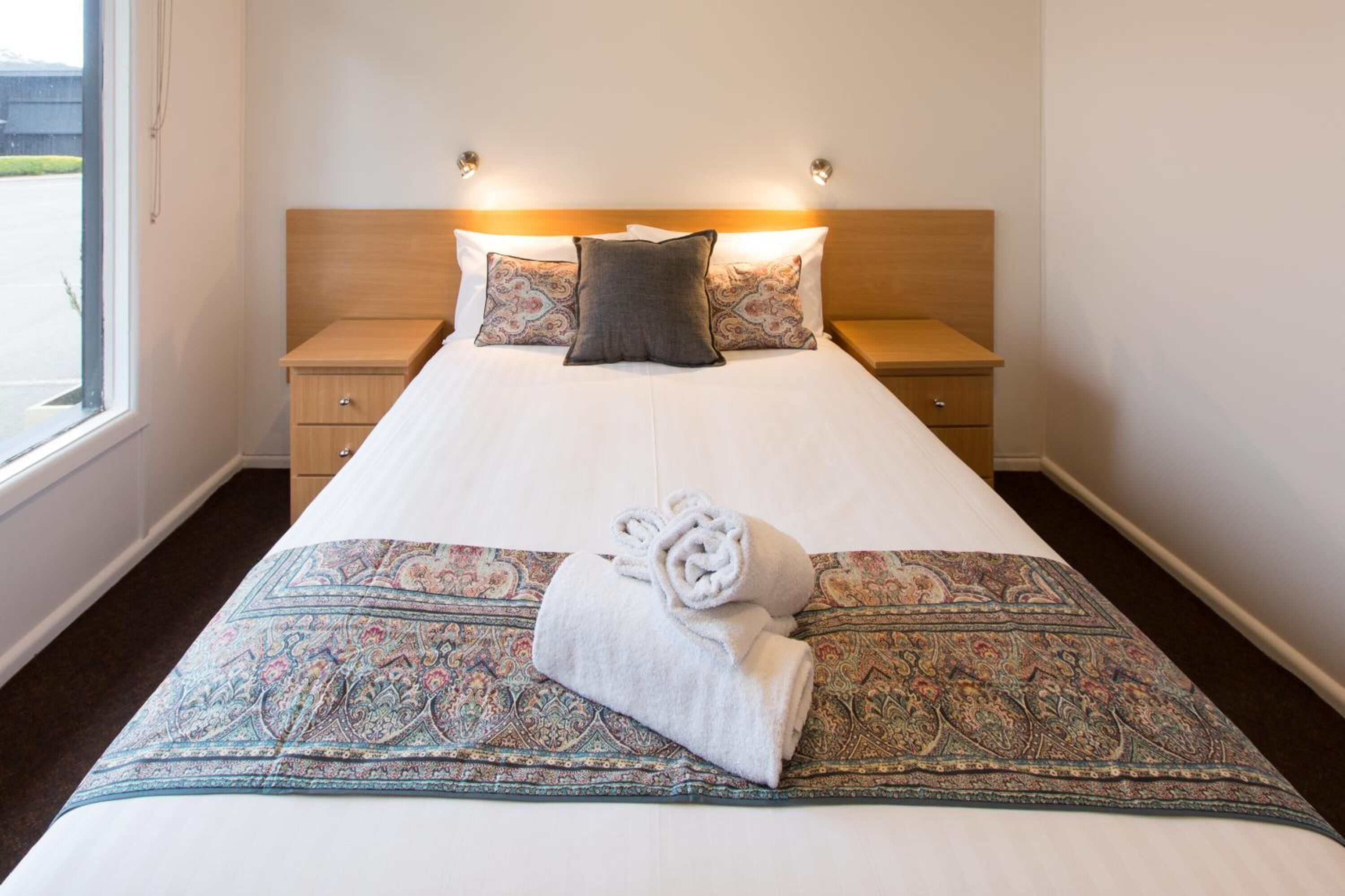 A neatly made bed with a patterned bedspread, two pillows, and a rolled towel on the bed.