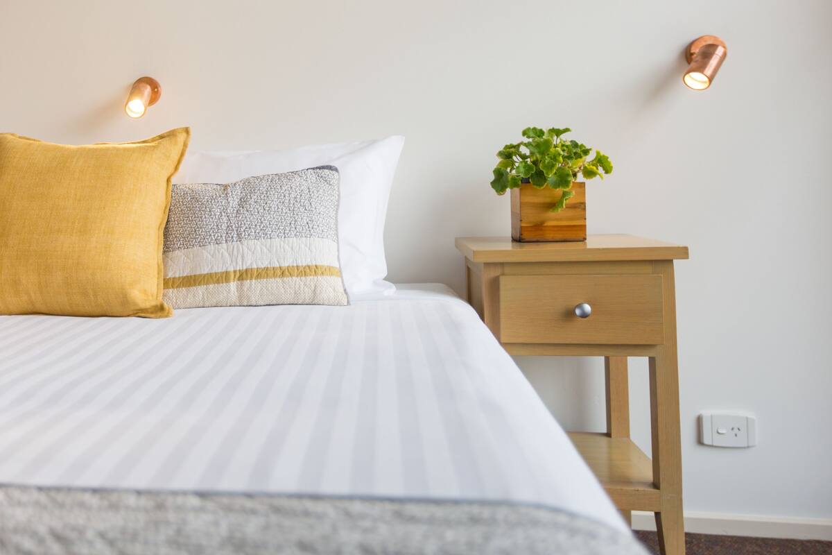 A neatly made bed with striped bedding, a yellow pillow, and a grey patterned pillow. There is a wooden nightstand with a potted plant and a wall-mounted light fixture.