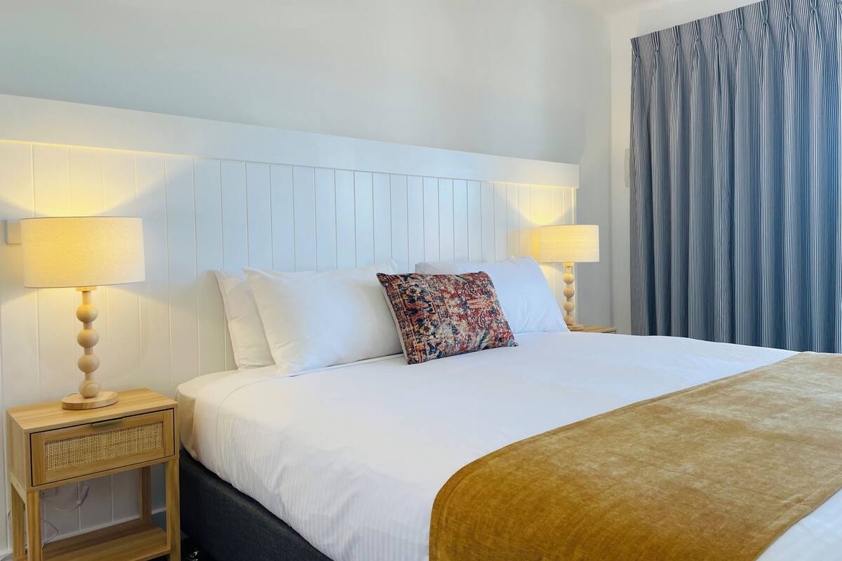 A hotel room with a large bed, two bedside tables with lamps, a patterned throw pillow, and a striped curtain.