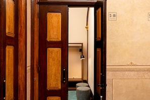 A wooden door with a brass handle, a small stool, and a lamp in a hallway with a beige wall and a door marked "3-B APARTMENT".