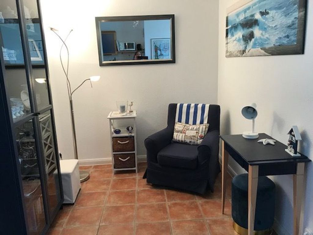 Un espacio habitable reducido con un sillón oscuro, una mesita con lámpara, un espejo y un cuadro en la pared.
