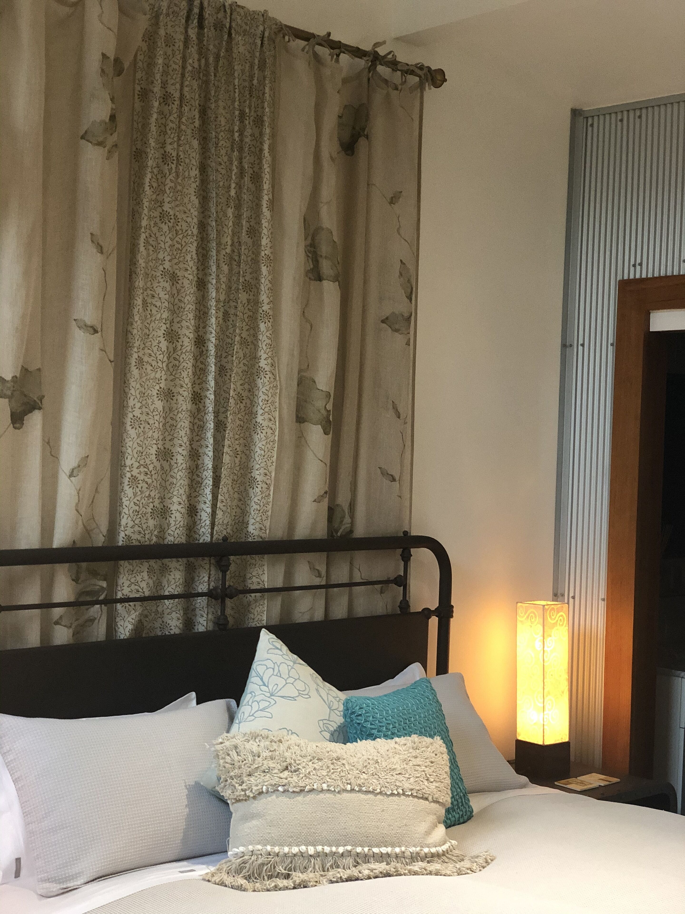 A bed with multiple pillows and a headboard, a bedside lamp, and a curtain with a floral pattern.