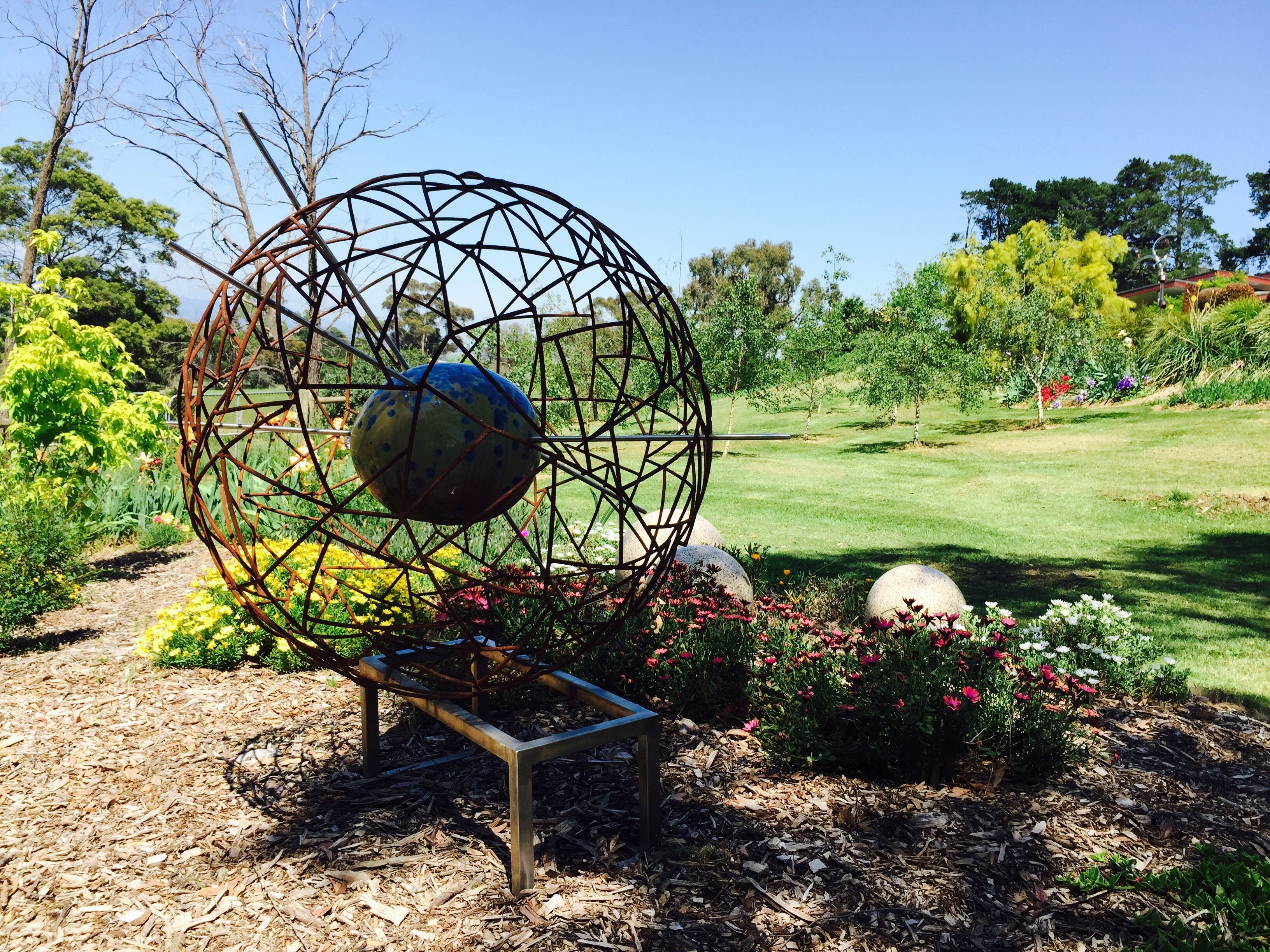 Un jardín con una escultura de metal, un banco y diferentes plantas.