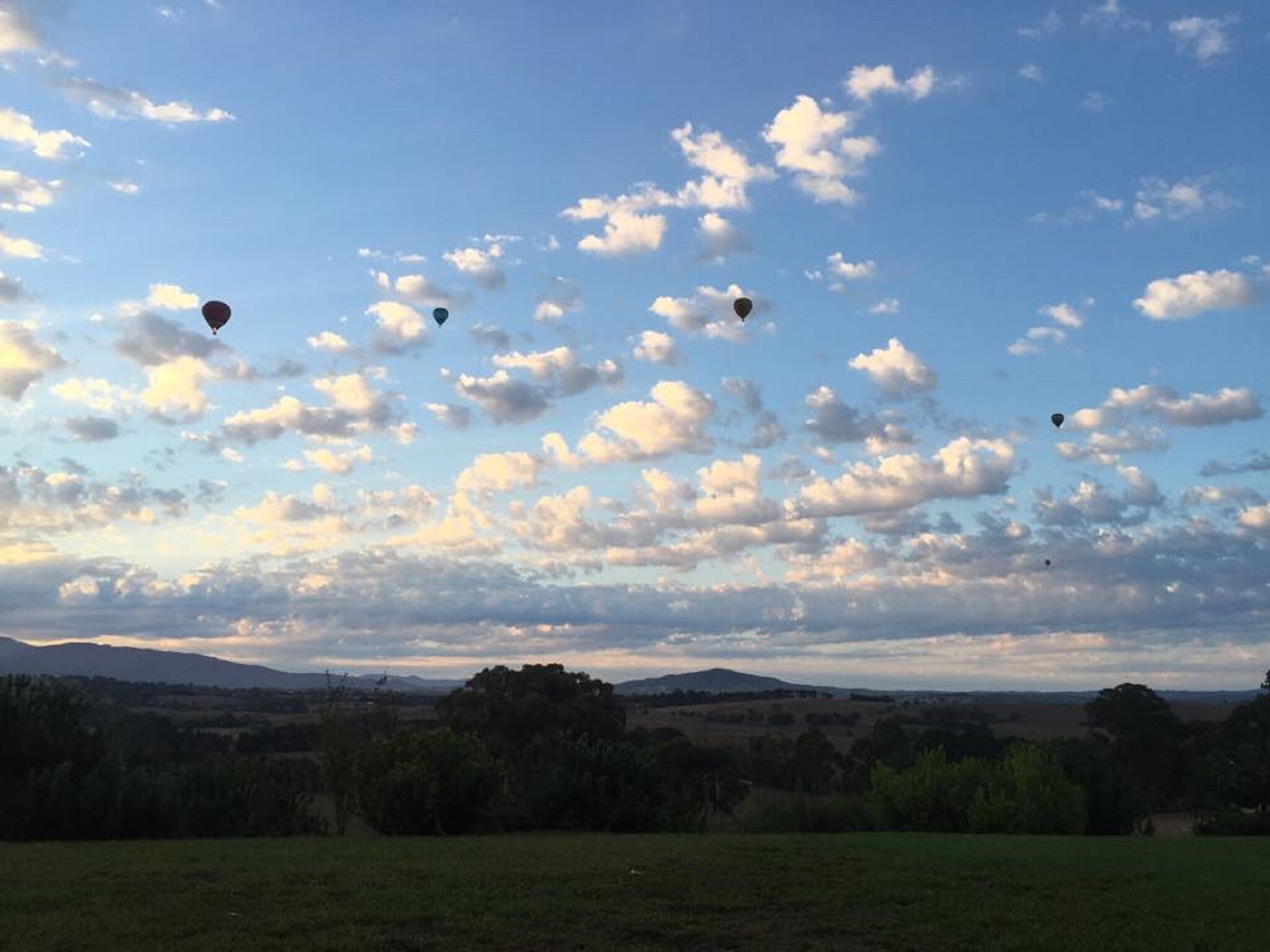 Un paysage avec de nombreux ballons à air chaud dans le ciel, un ciel dégagé parsemé de quelques nuages, et une vue lointaine sur des collines et des arbres.