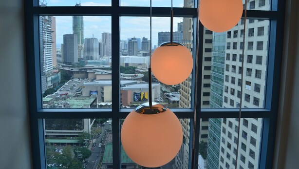 A view through a window with three hanging spherical light fixtures, overlooking a cityscape with high-rise buildings and construction cranes. - Eton Parkview (Makati)