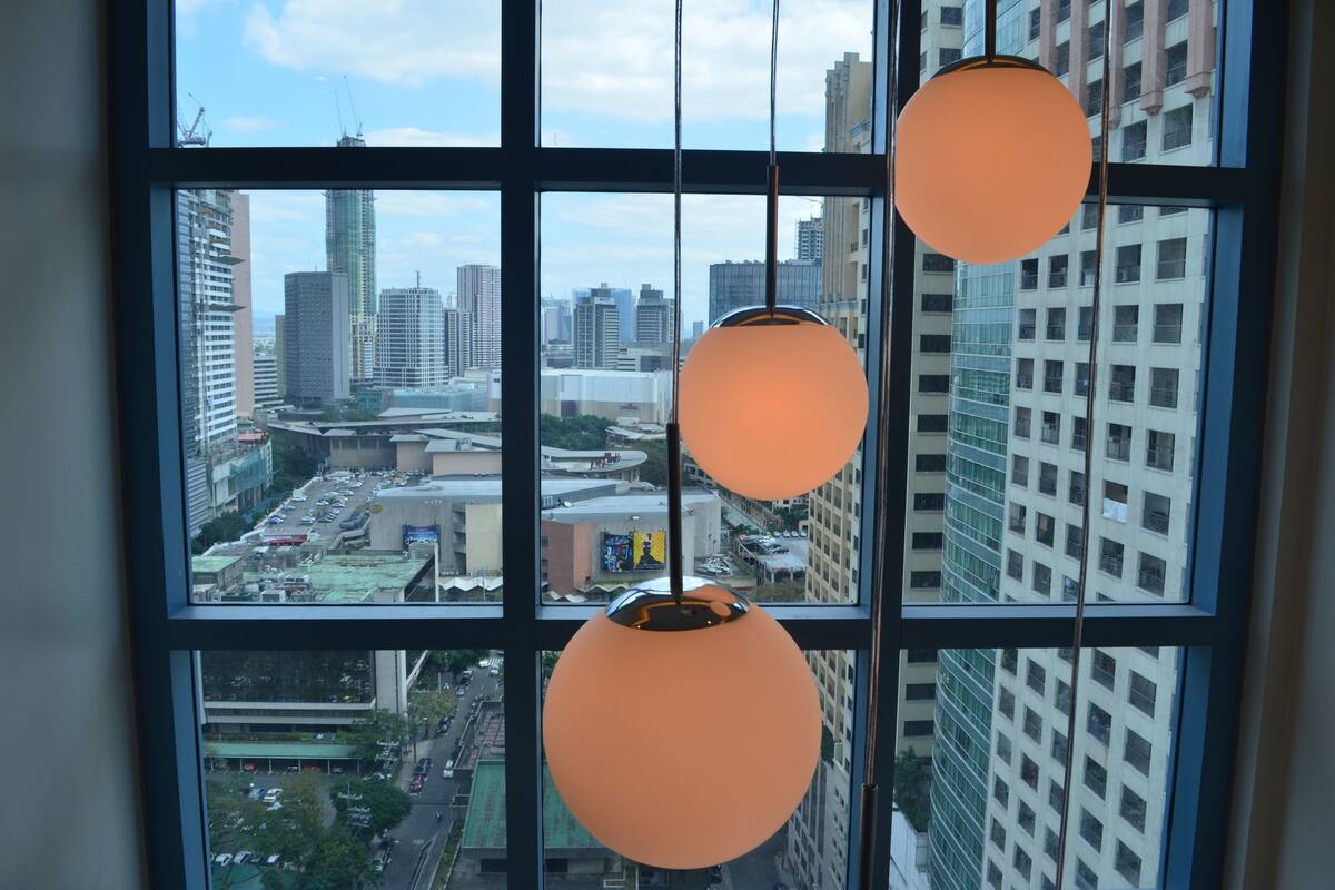 A view through a window with three hanging spherical light fixtures, overlooking a cityscape with high-rise buildings and construction cranes.