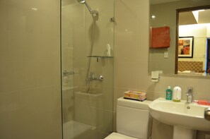 A bathroom with a glass-enclosed shower, a white sink with toiletries, and a mirror reflecting a room with a red towel and a framed picture.