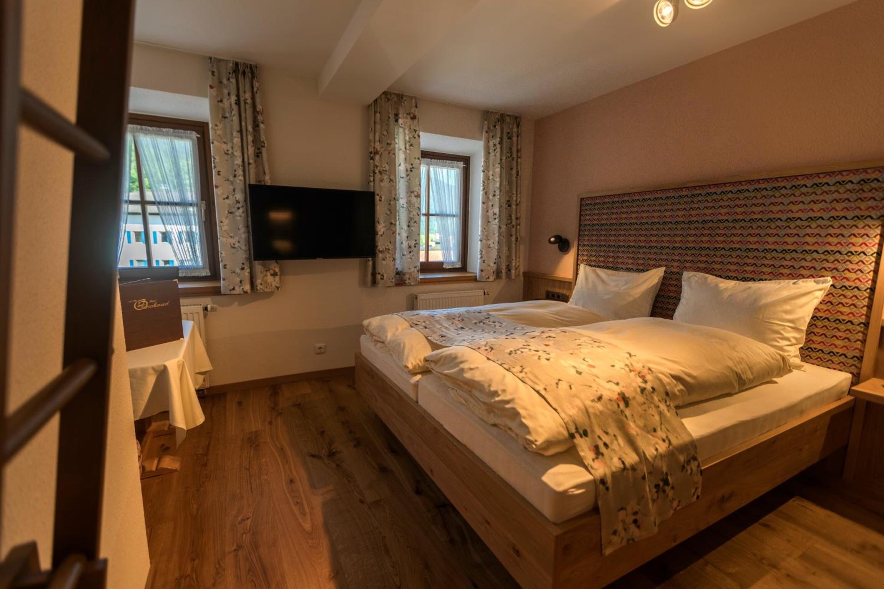 Ein Schlafzimmer mit einem hölzernen Bett, zwei mit Vorhängen versehenen Fenstern, einem Flachbildfernseher und einem Nachttisch.