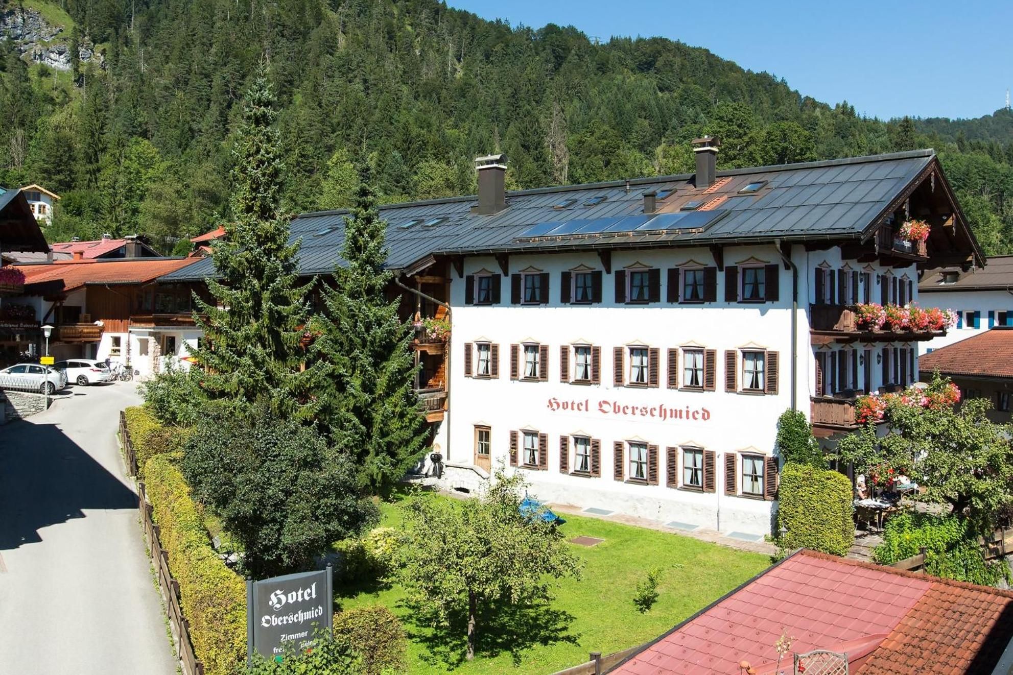 A hotel with a white facade and brown shutters, surrounded by greenery and a mountain backdrop.