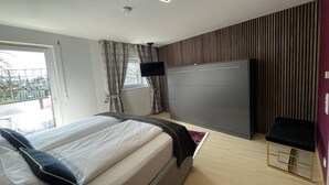 A modern bedroom with a bed, a TV mounted on the wall, and a window with curtains. - Design Apartment Catch Me If You Can (Freiburg im Breisgau)