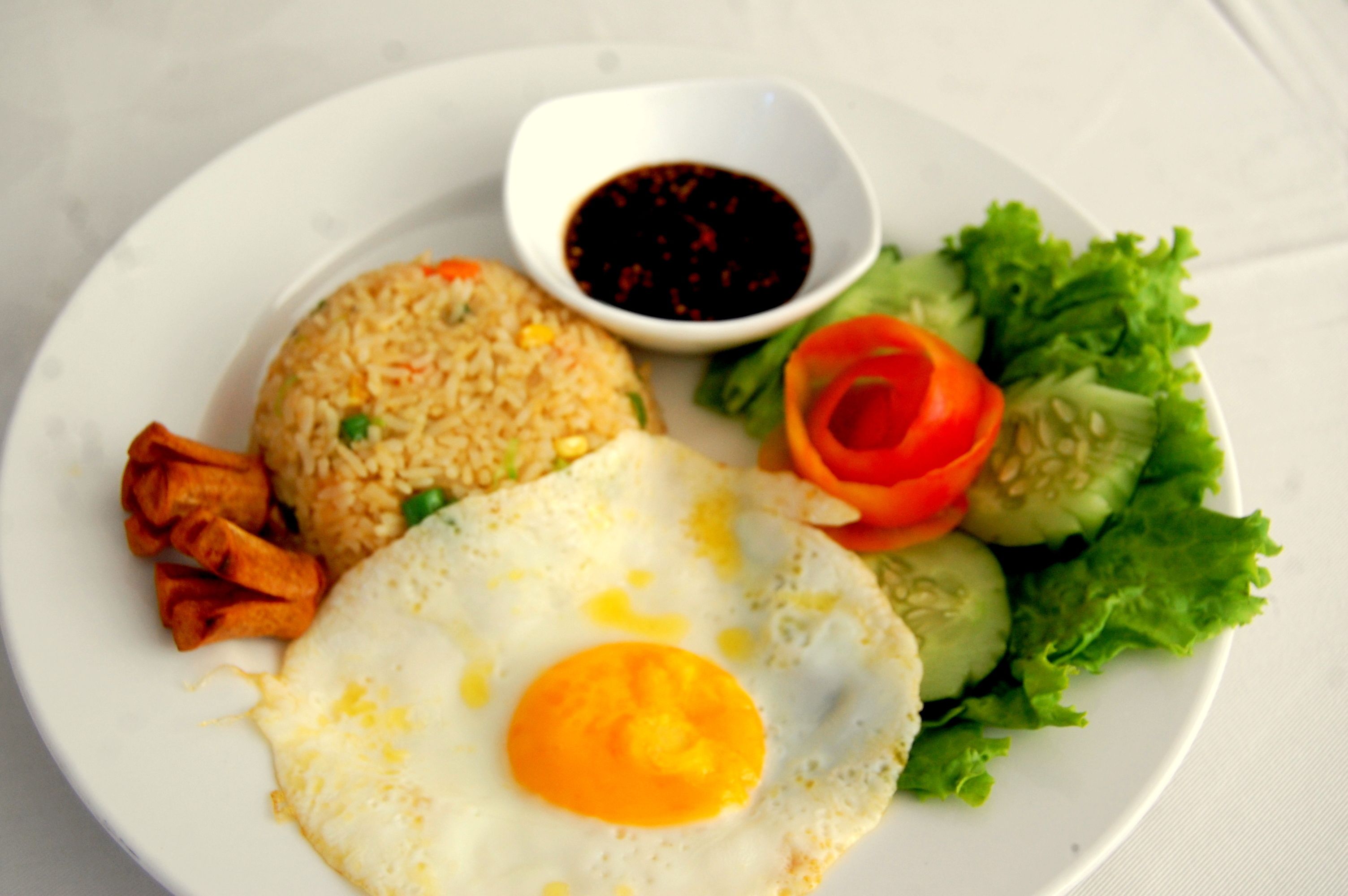 A plate with fried rice, a fried egg, a slice of tomato, cucumber, lettuce, and a small bowl of sauce.