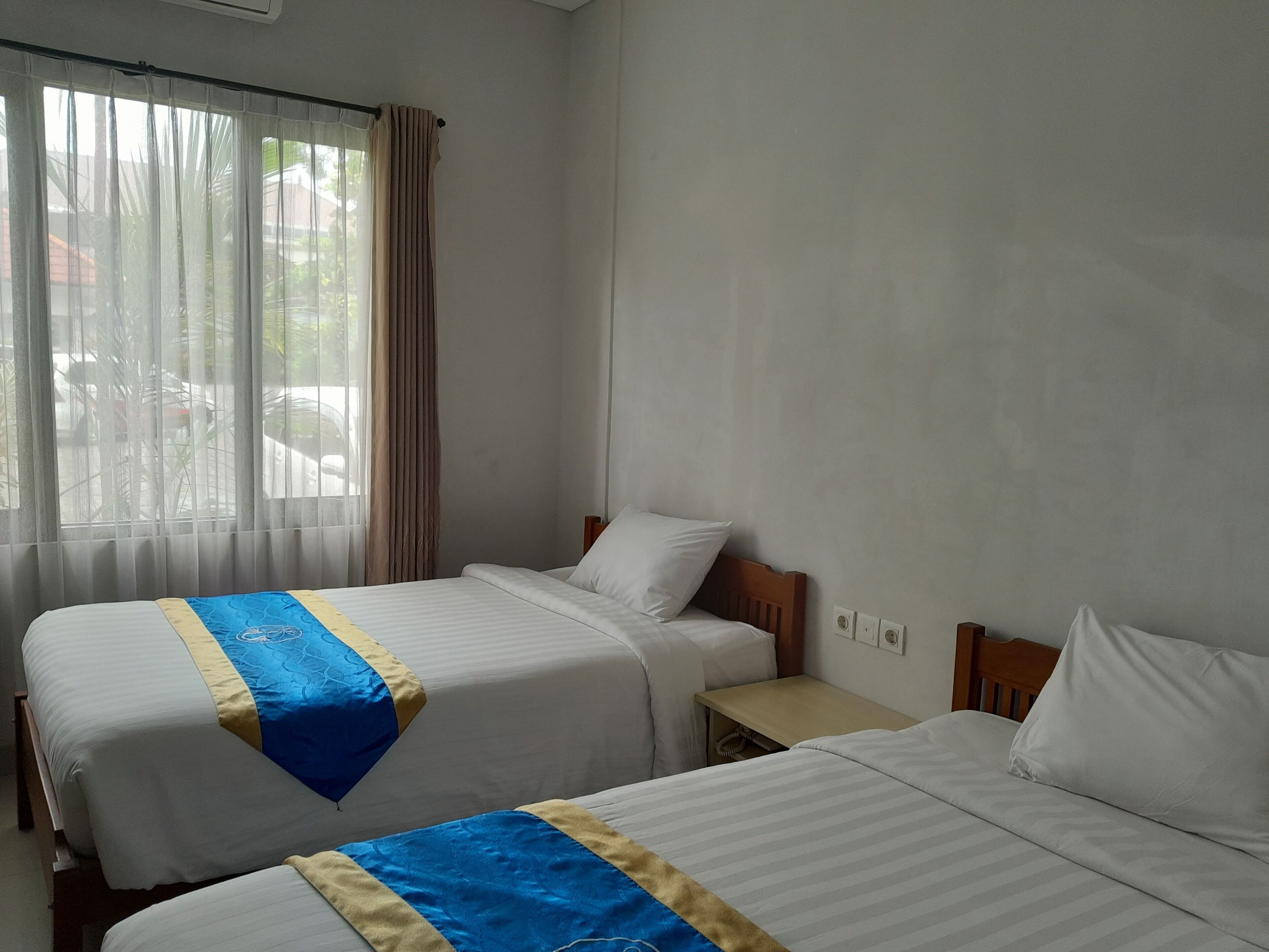Two single beds with white linens and blue and yellow decorative borders, a small bedside table, and a window with sheer curtains.
