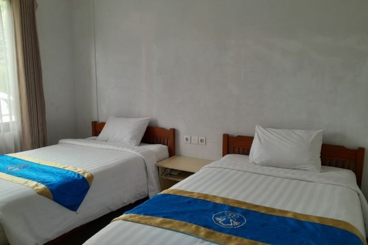Una habitación de hotel con dos camas, cada una con ropa de cama blanca y almohadas decorativas azules y doradas. Hay una mesita de noche de madera entre las camas.