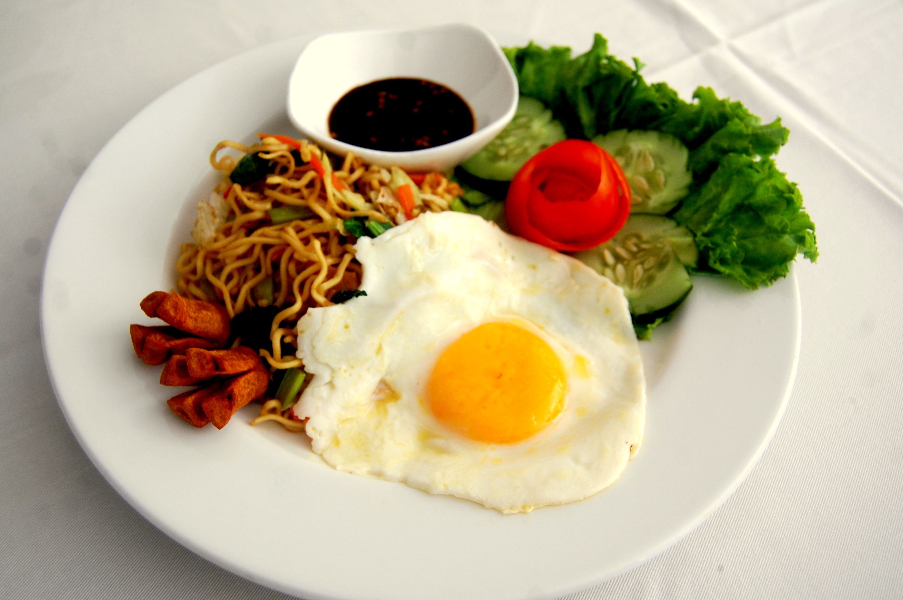 A plate with a fried egg, noodles, vegetables, and a small bowl of sauce.