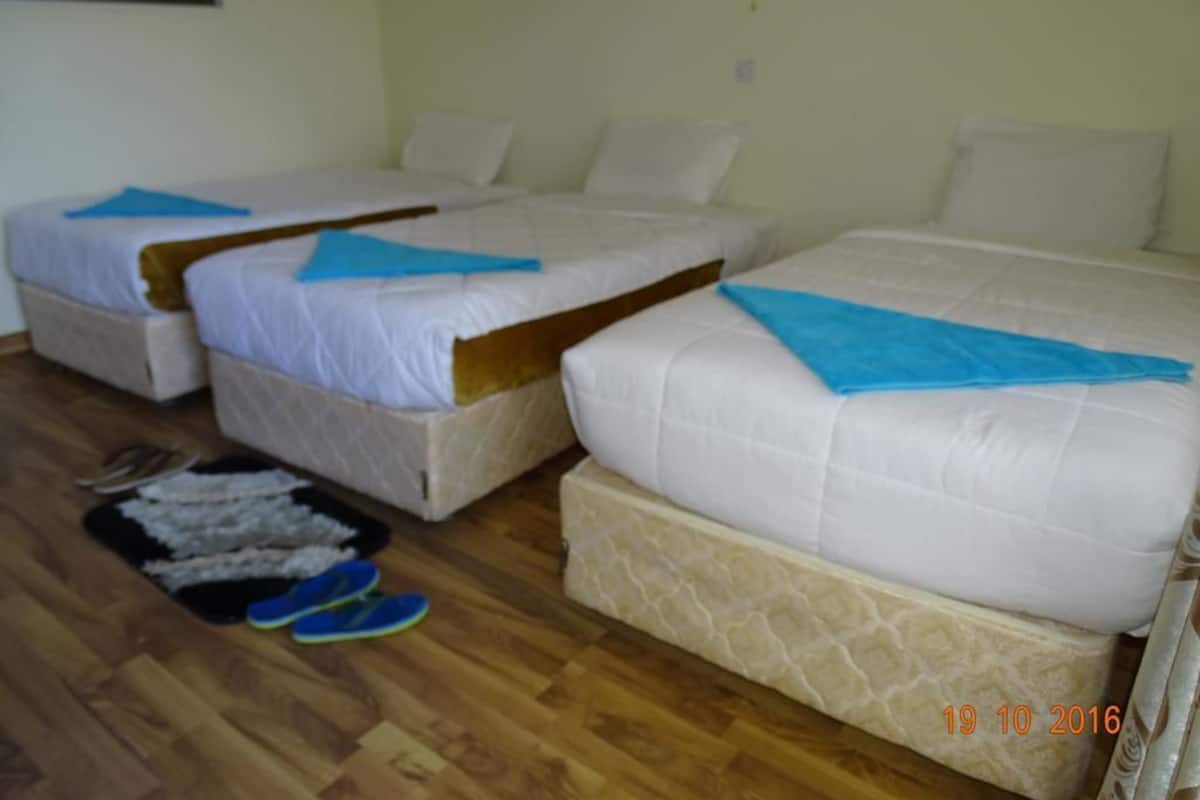 A room with three single beds, each with a blue towel and white bedding. There is a pair of blue slippers on the floor.