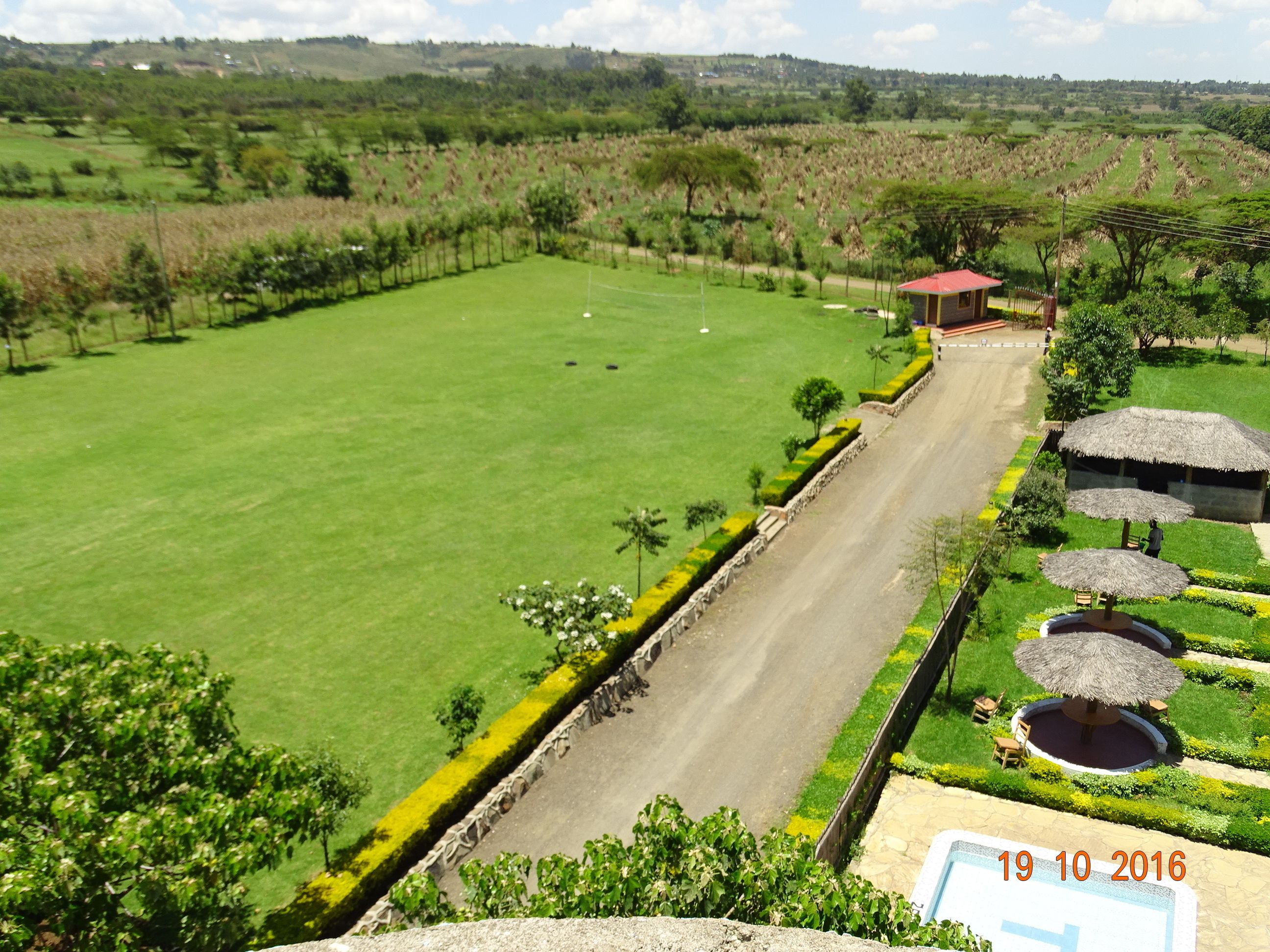 A large green field with a paved path, a small building with a red roof, and thatched umbrellas.
